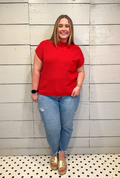 Woman wearing a red shirt and blue jeans standing against a light gray wall.
