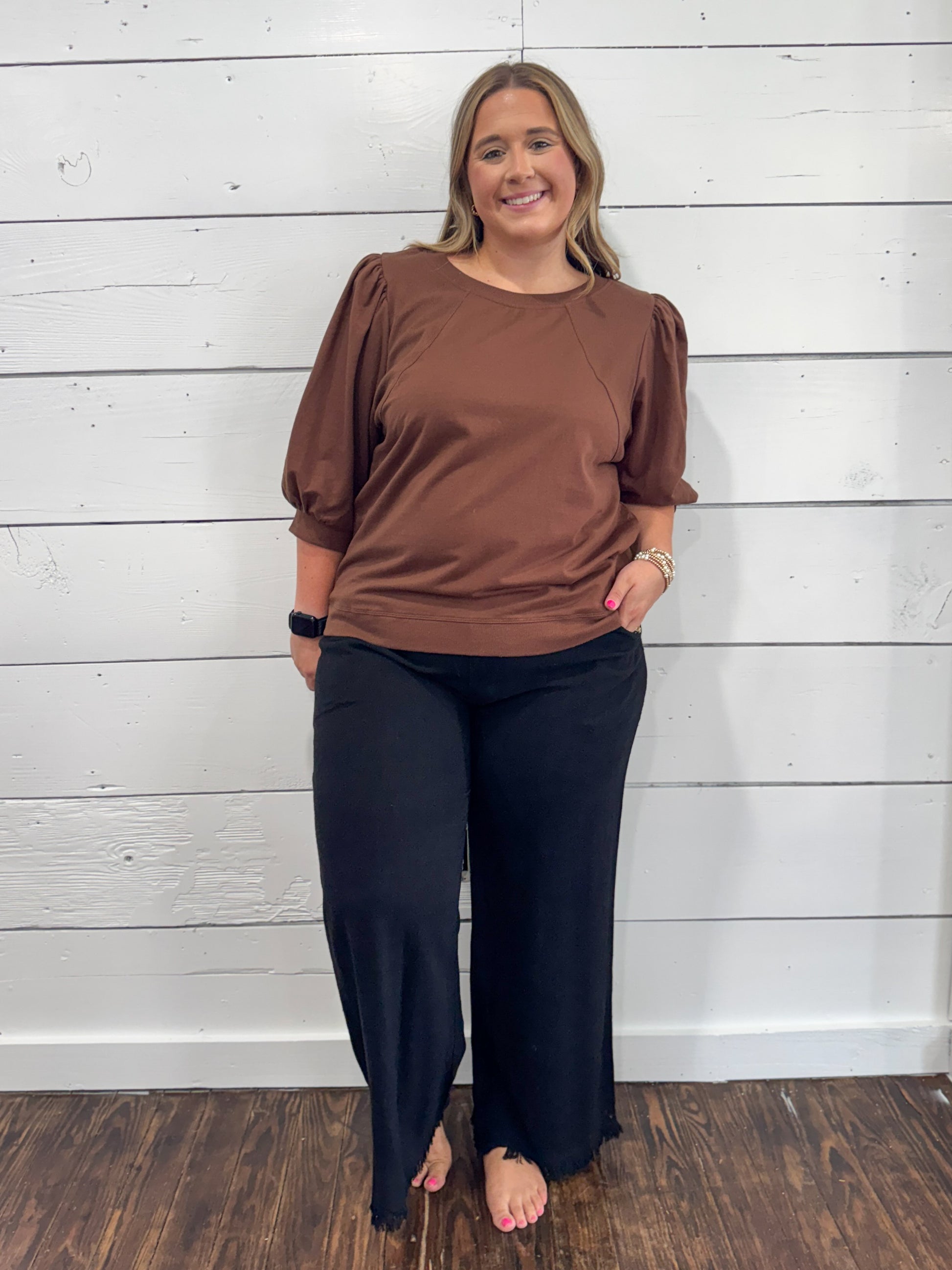 A woman modeling a brown short-sleeve top with puffed sleeves and black wide-leg pants, standing against a white shiplap wall on a dark wood floor.