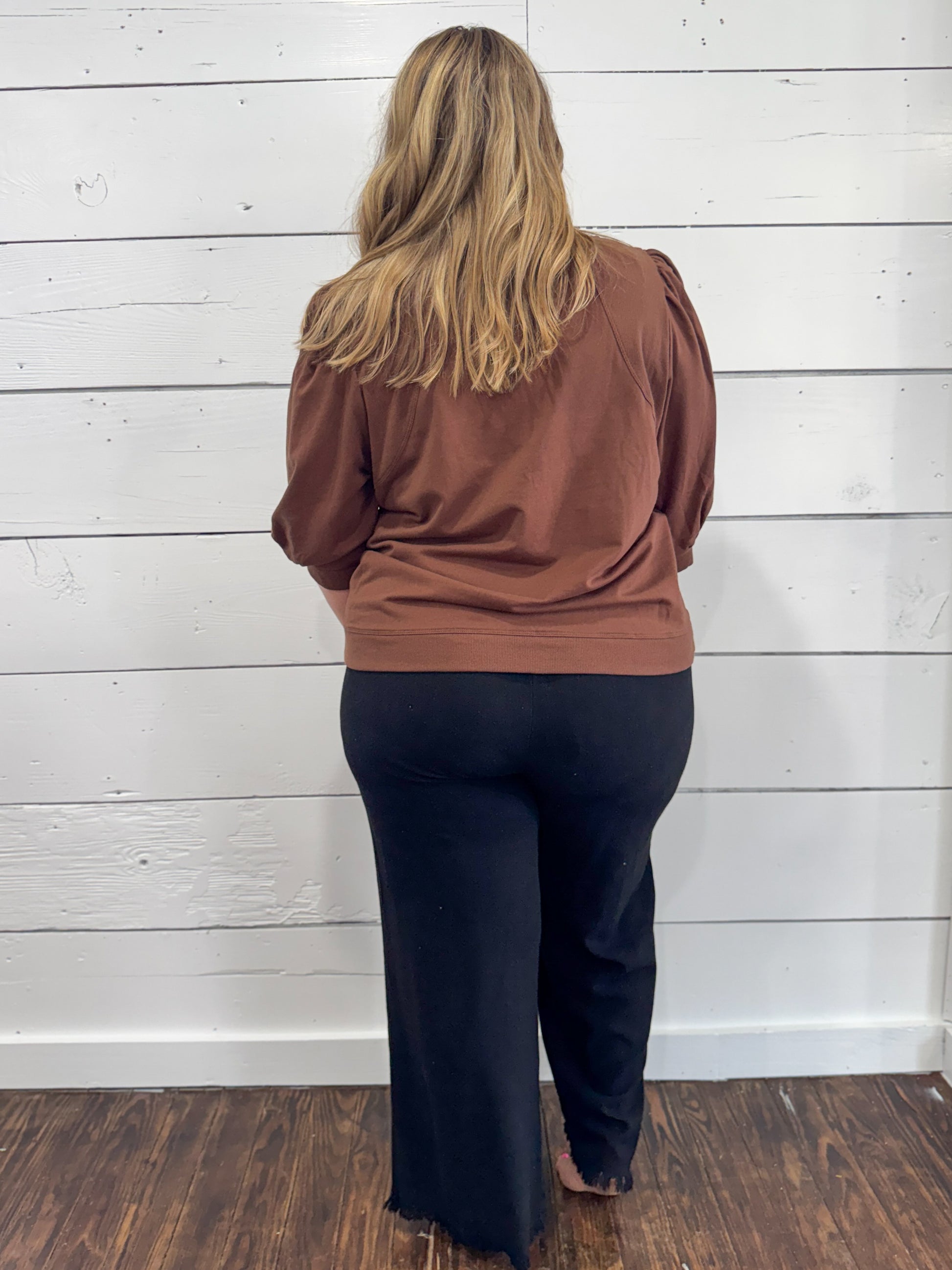 A woman viewed from behind wearing a rust-brown long-sleeve top and dark navy wide-leg pants, standing against a white wood-paneled wall on a dark wood floor.