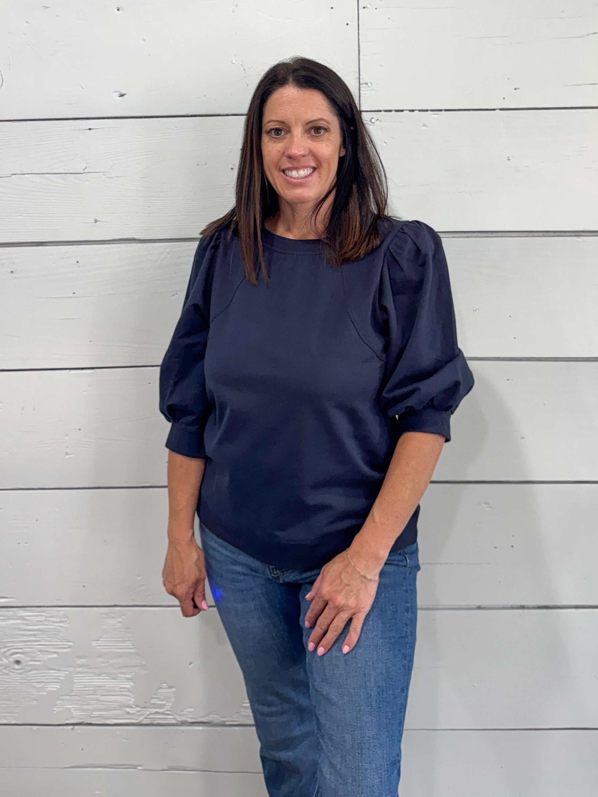 A woman wearing a navy blue long-sleeve blouse with puffed shoulders and blue jeans, standing against a white shiplap wall.