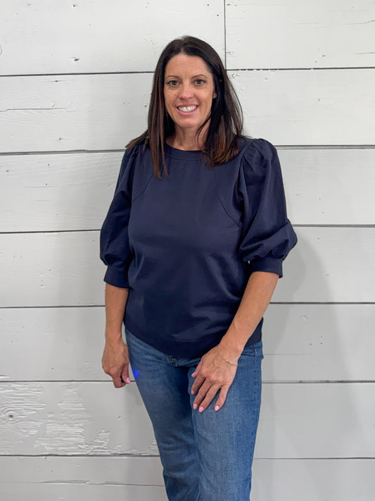 A woman wearing a navy blue long-sleeve blouse with puffed shoulders and blue jeans, standing against a white shiplap wall.