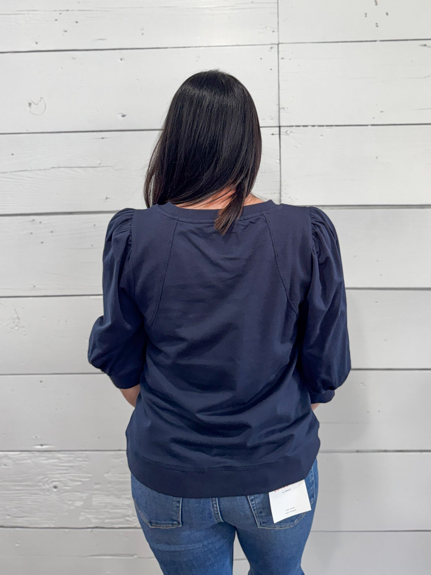 A woman viewed from behind wearing a navy blue long-sleeve top with puffed sleeves and blue jeans, standing against a white wood plank wall.
