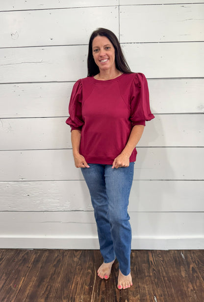 A woman wearing a magenta puff-sleeve top and blue jeans, standing barefoot on a dark wood floor against a white shiplap wall.