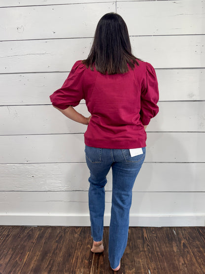 A woman viewed from behind wearing a magenta puff-sleeve top and blue jeans, standing against a white wood-paneled wall on a dark wood floor.