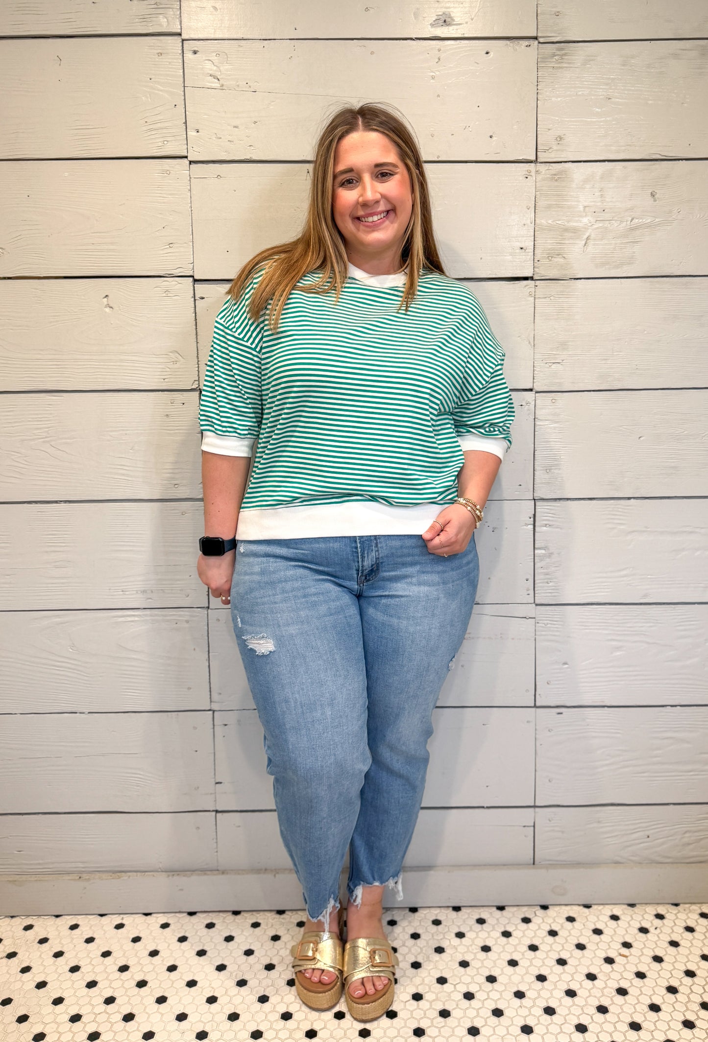 Woman wearing a green striped shirt and blue jeans standing against a light wooden paneled wall.