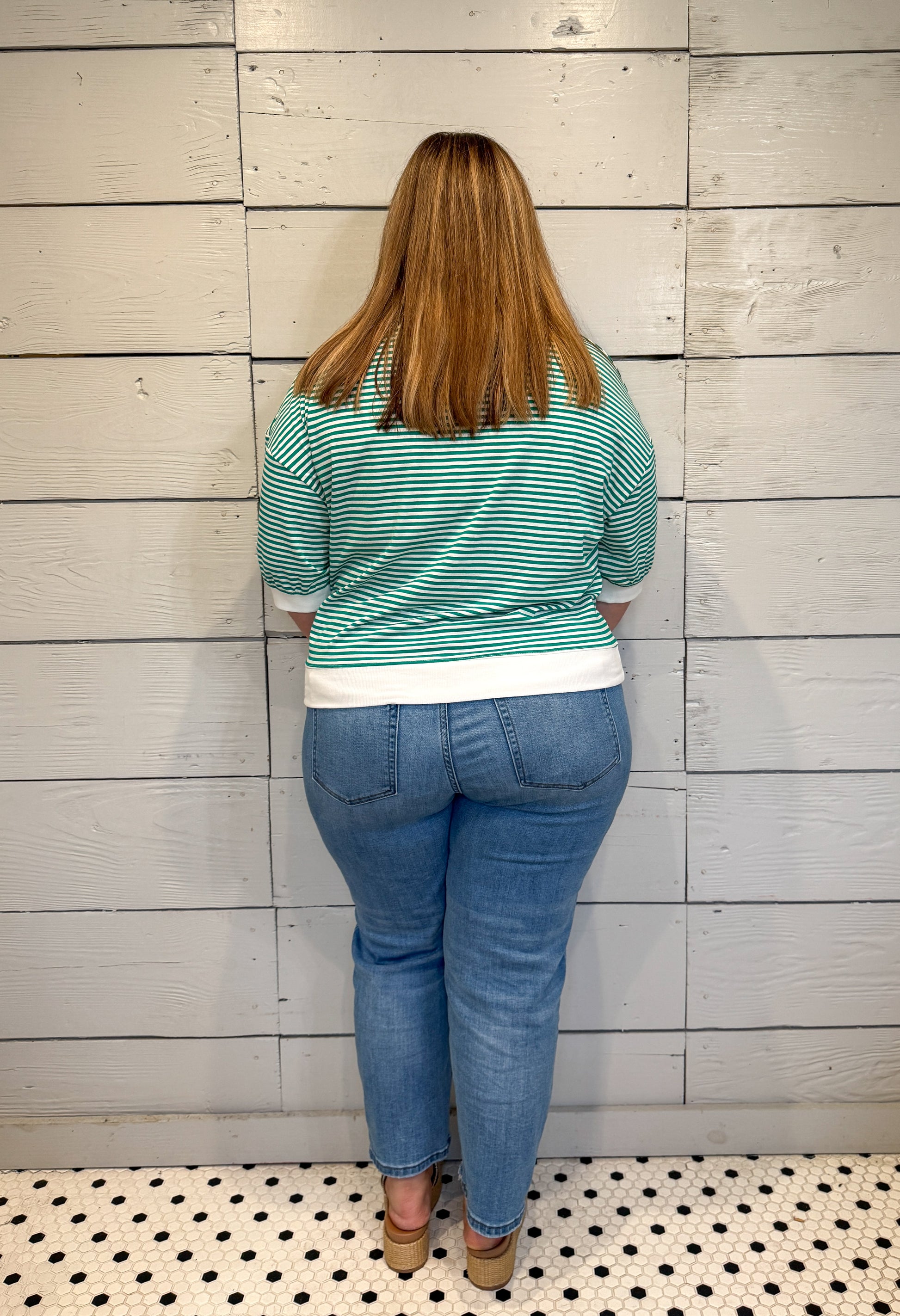 Person wearing a green striped shirt and blue jeans standing against a light wooden paneled wall.