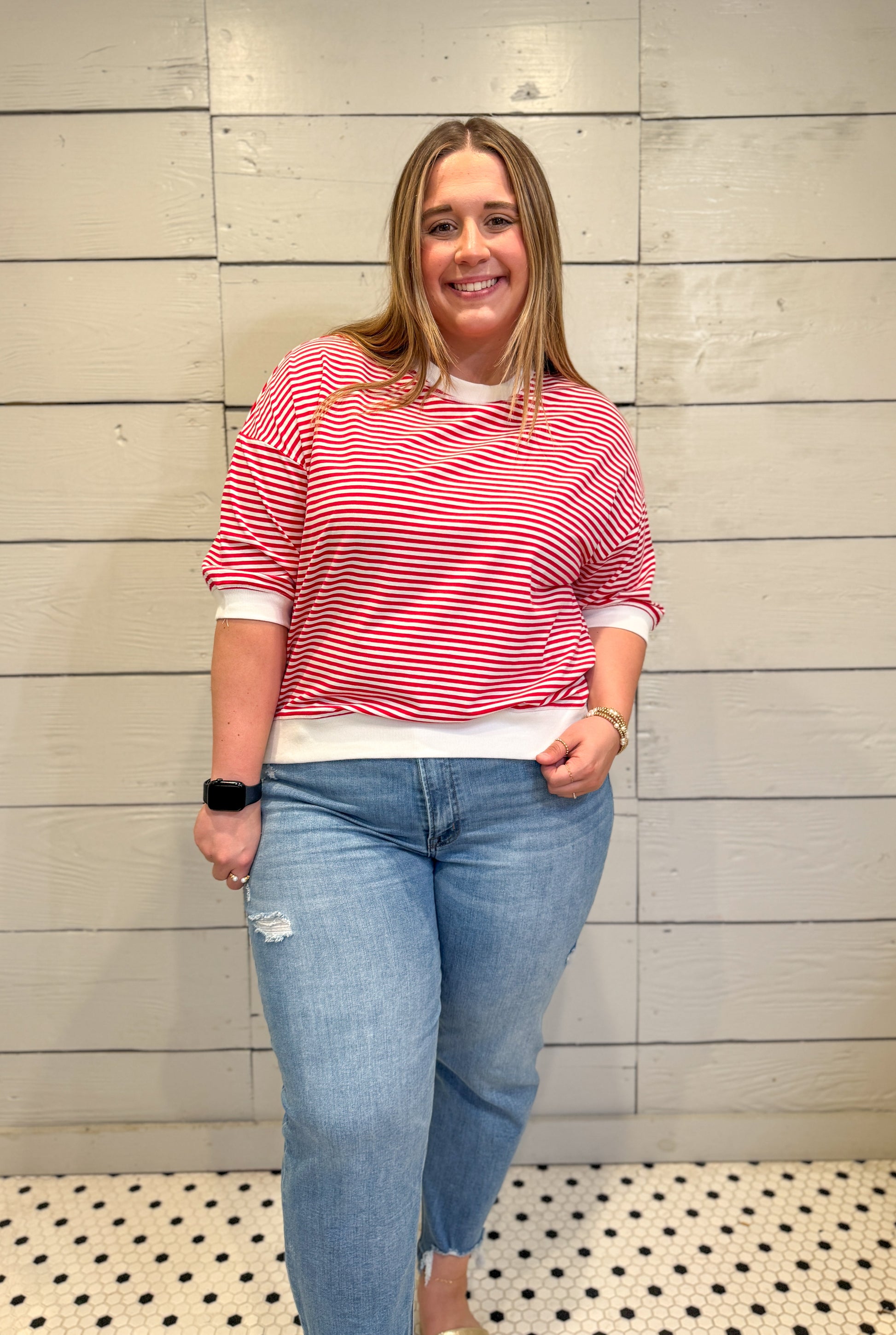 Woman wearing a red and white striped shirt and blue jeans standing against a wooden paneled wall.