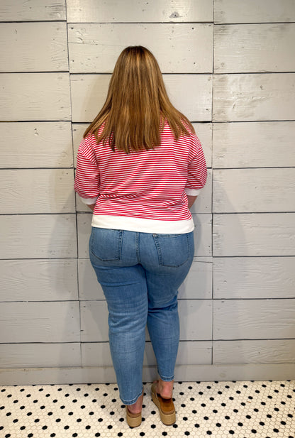 Person wearing a red and white striped shirt and blue jeans standing against a wooden paneled wall.