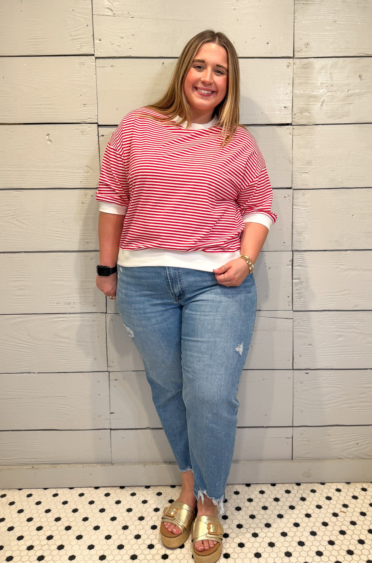 Woman wearing a red and white striped shirt, blue jeans, and gold sandals standing against a light wooden paneled wall.