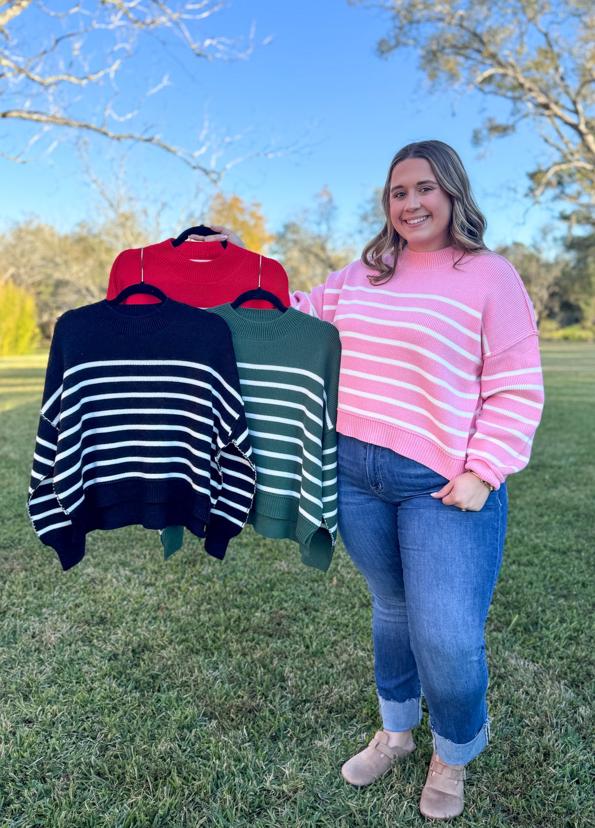 Woman standing outdoors with three sweaters displayed behind her
