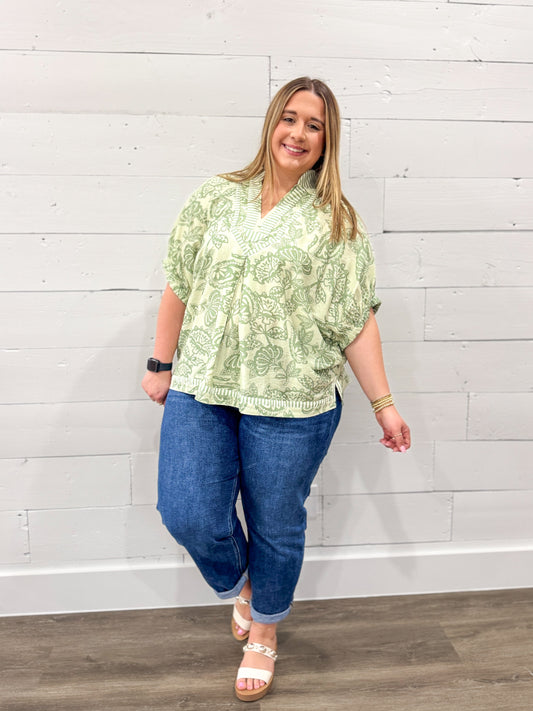 Woman wearing a green patterned blouse and blue jeans against a white wooden panel background