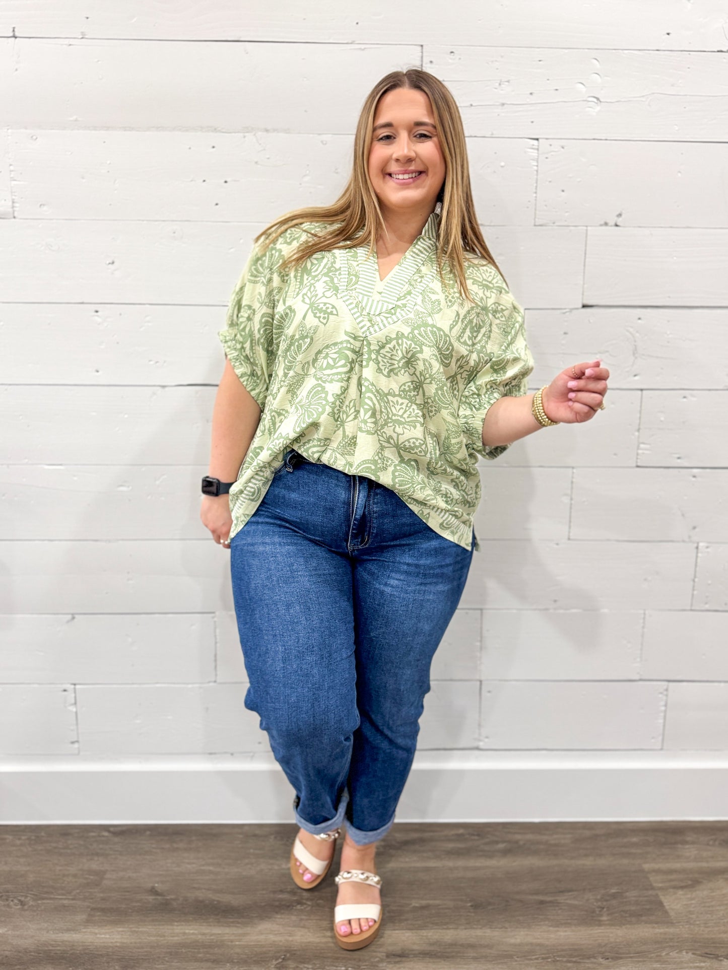 Woman wearing a green patterned blouse and blue jeans against a white brick wall.
