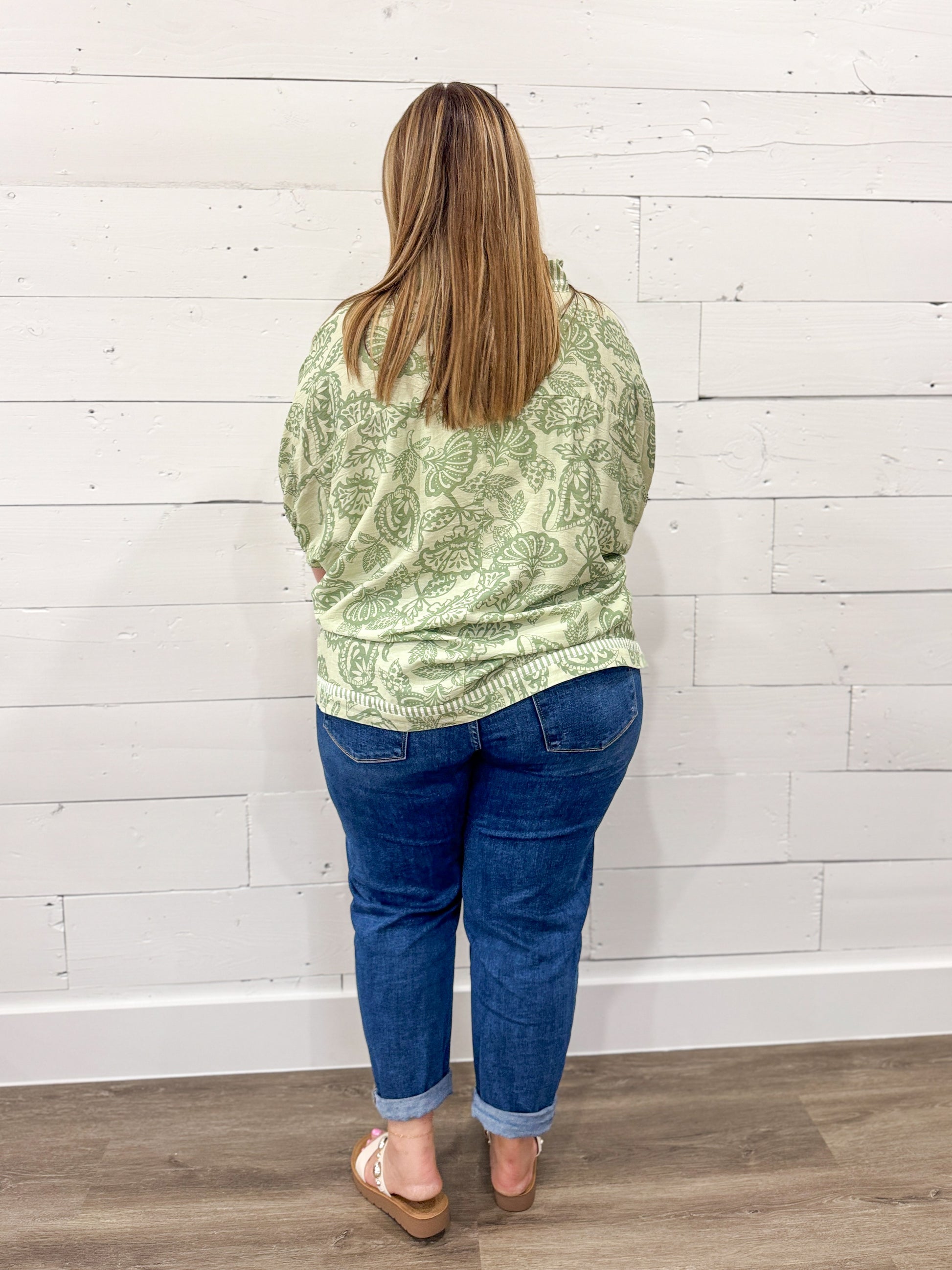 Person wearing a green patterned top and blue jeans standing against a white brick wall.