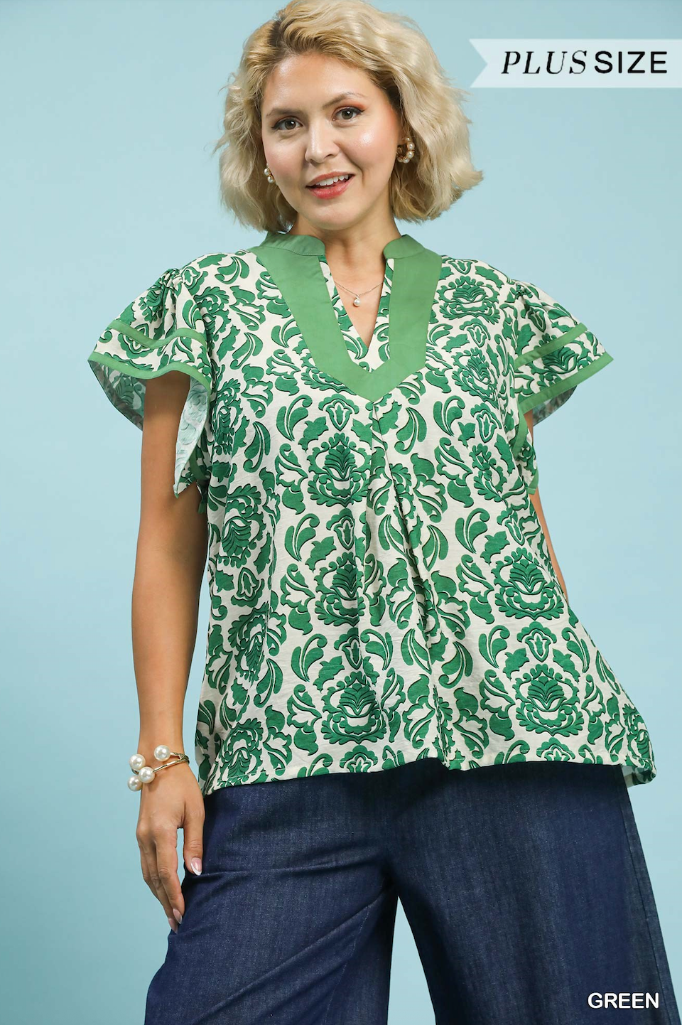 Woman wearing a green and white patterned blouse with blue pants on a light blue background.