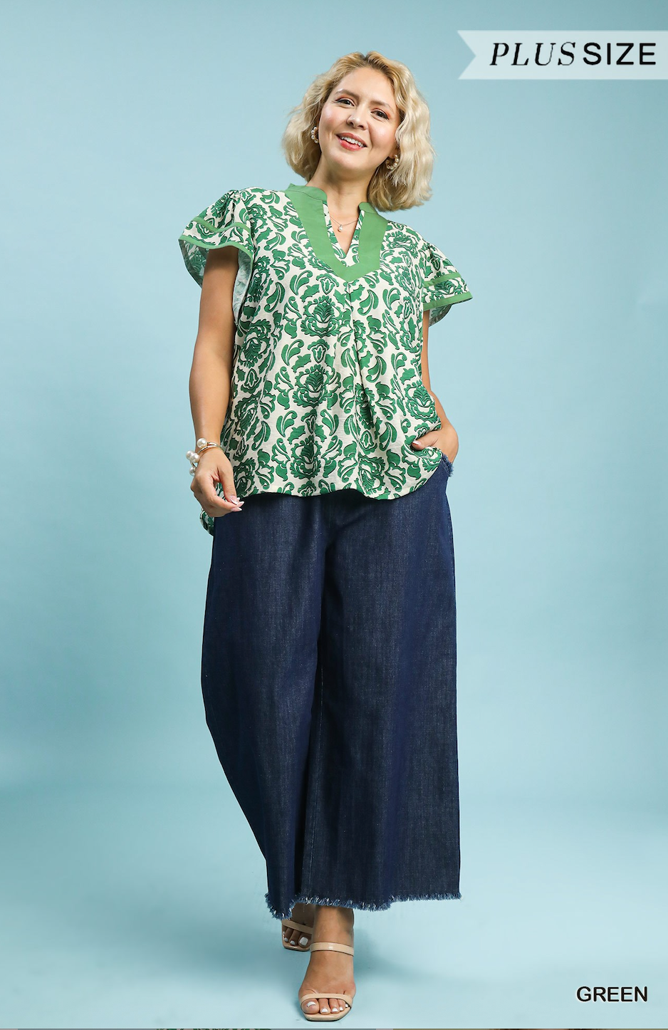 Woman wearing a green patterned blouse and navy pants on a light blue background