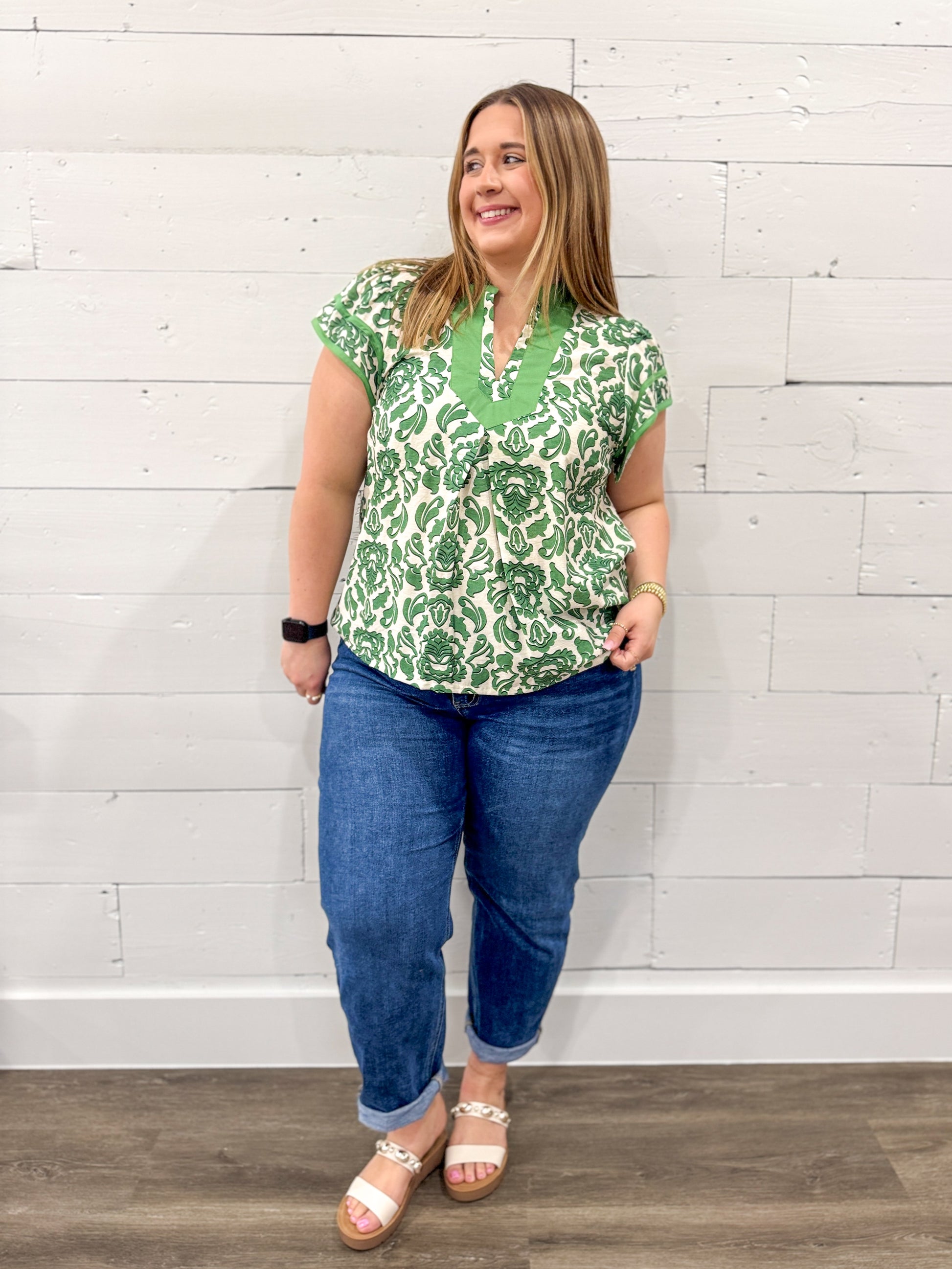 Woman wearing a green patterned top and blue jeans against a white brick wall.