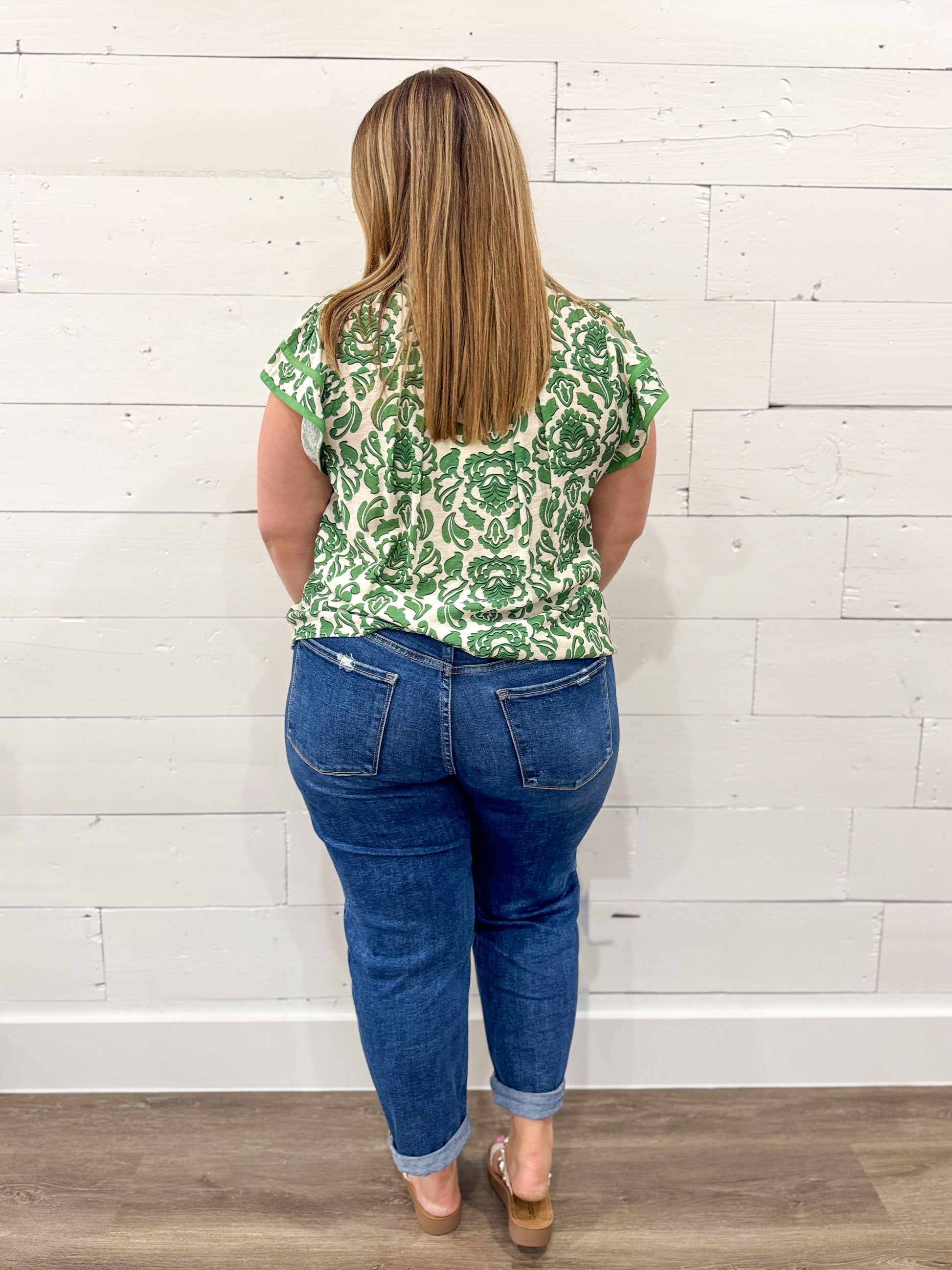 Woman wearing a green patterned top and blue jeans standing against a white brick wall.