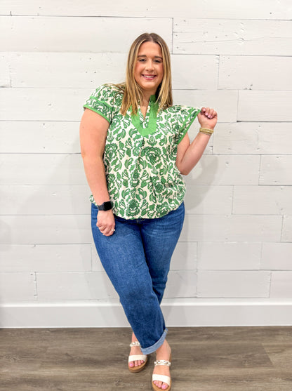 Woman wearing a green patterned top and blue jeans against a white brick wall.