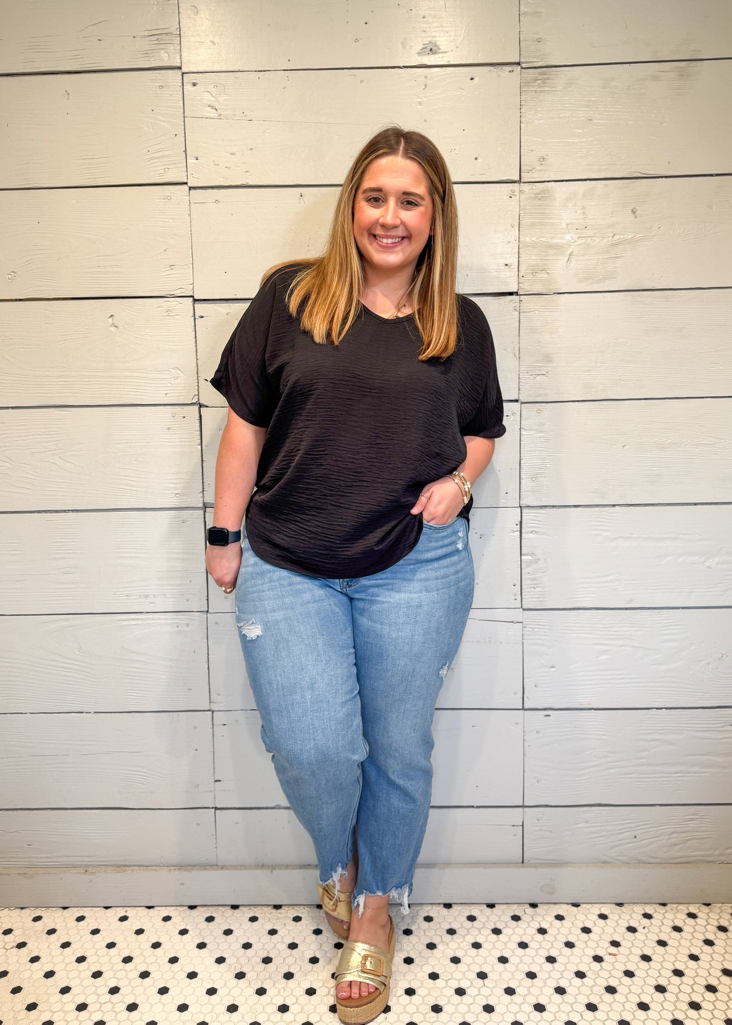 Woman wearing a black t-shirt and blue jeans standing against a light wooden paneled wall.