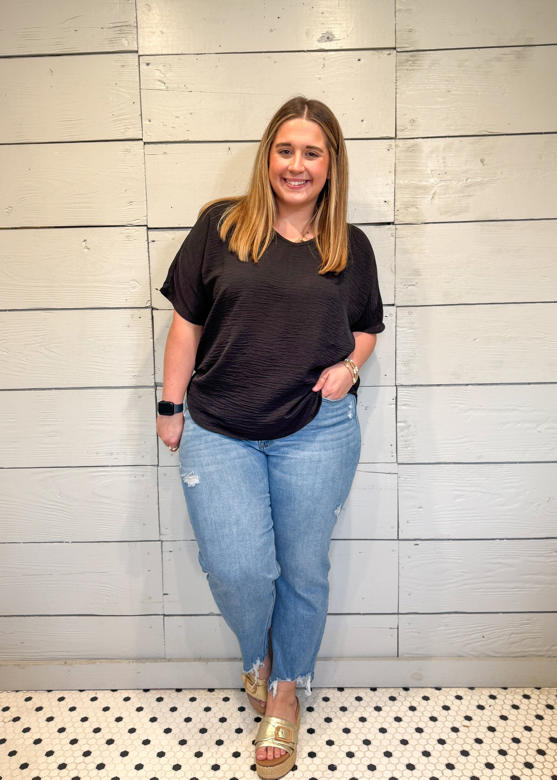 Woman wearing a black t-shirt and blue jeans standing against a light wooden paneled wall.