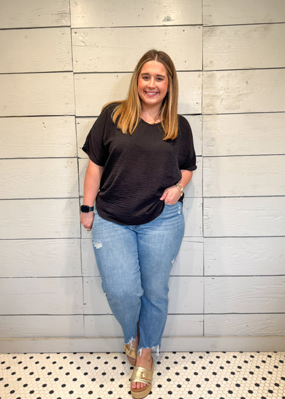 Woman wearing a black t-shirt and blue jeans standing against a light wooden paneled wall.