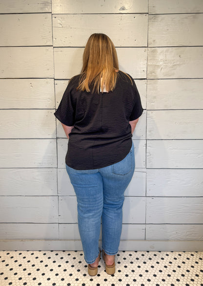 Person wearing a black t-shirt and blue jeans standing against a light wooden panel wall.