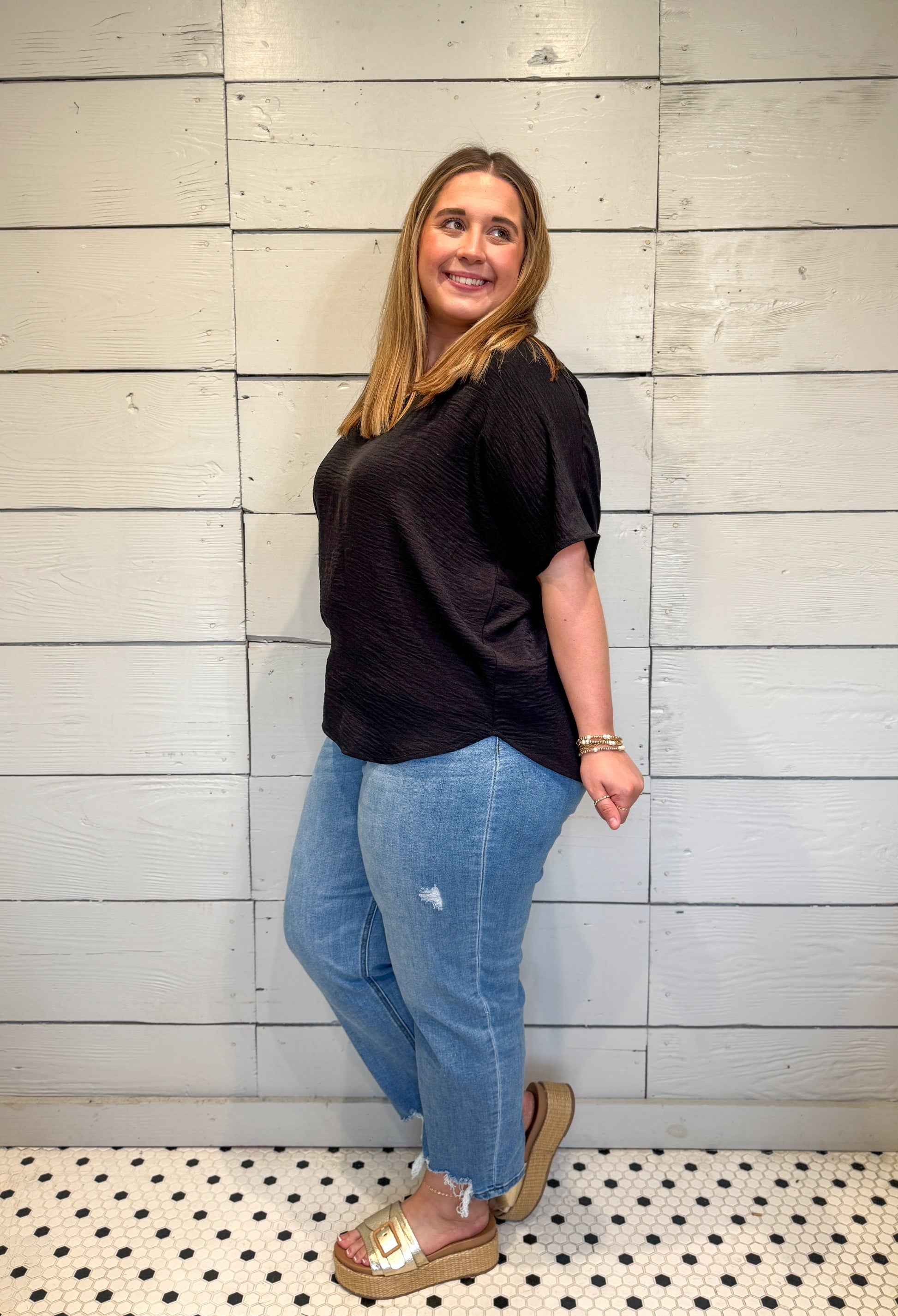 Woman wearing a black top and blue jeans standing against a light wooden panel wall.