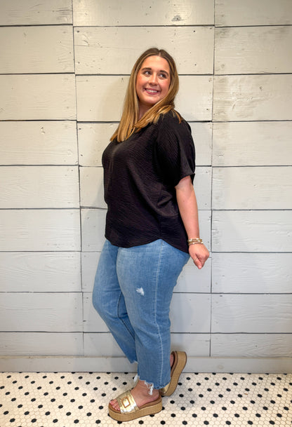 Woman wearing a black top and blue jeans standing against a light wooden panel wall.