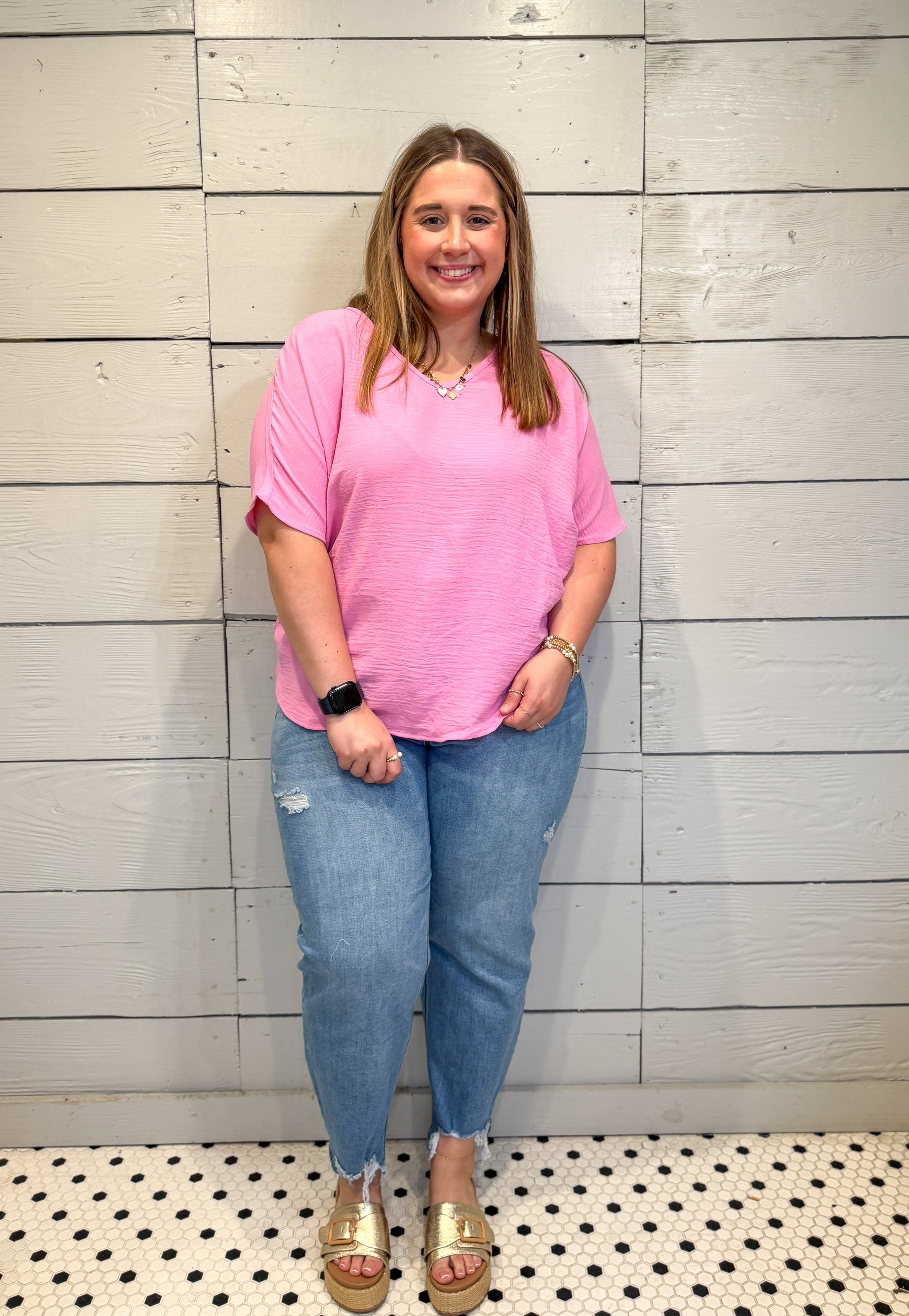 Woman wearing a pink shirt and blue jeans standing against a light gray wall.