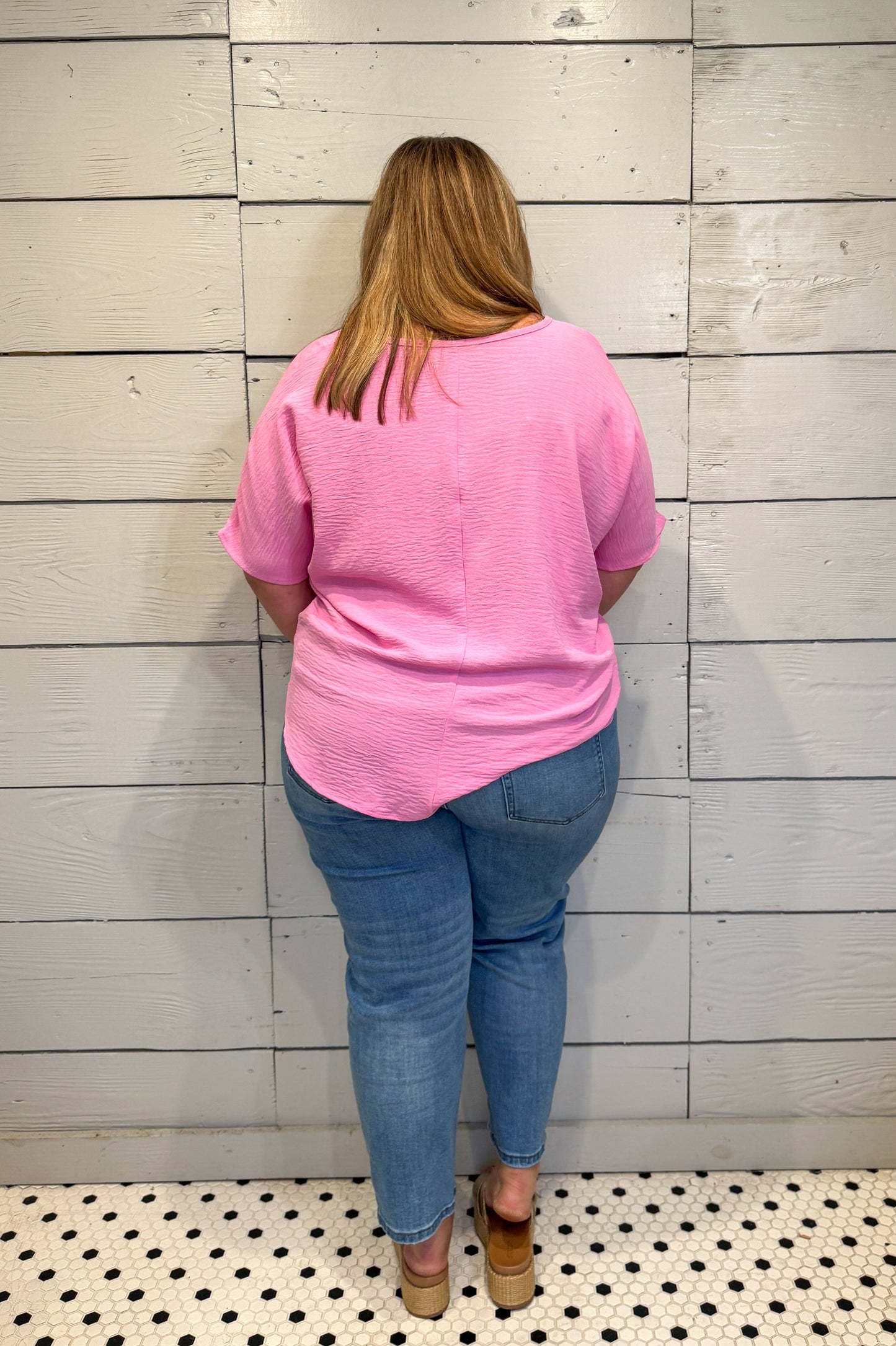 Person wearing a pink shirt and blue jeans standing against a light wooden paneled wall.