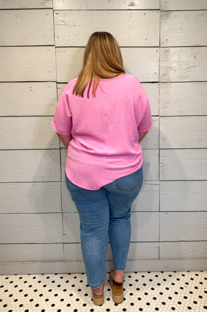Person wearing a pink shirt and blue jeans standing against a light wooden paneled wall.