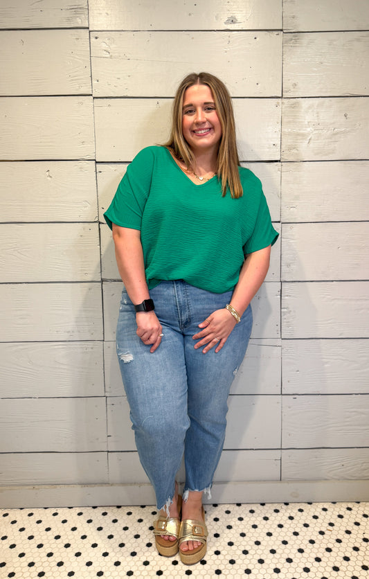 Woman wearing a green top and blue jeans standing against a light wooden paneled wall.