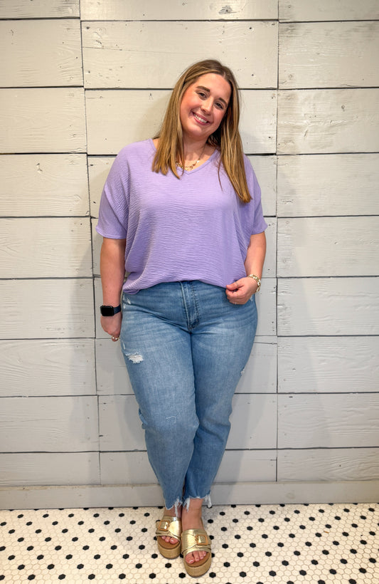 Woman wearing a purple top and blue jeans standing against a light gray wall.