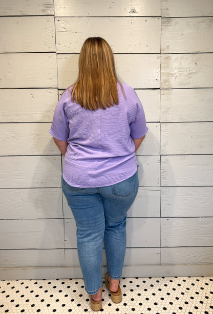 Person wearing a purple shirt and blue jeans standing against a light wooden paneled wall.
