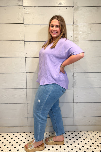 Woman wearing a light purple t-shirt and blue jeans standing against a light wooden paneled wall.