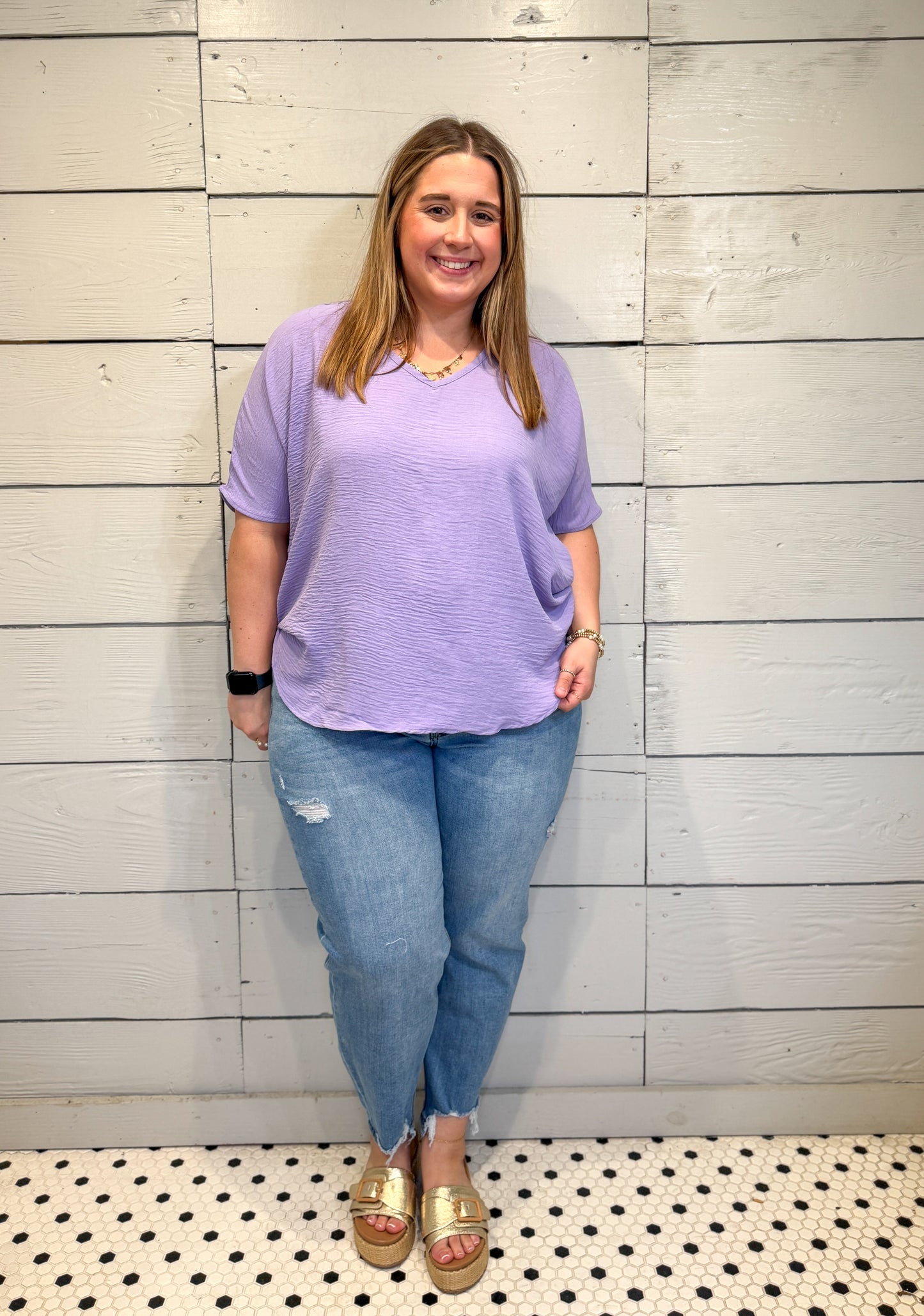 Woman wearing a purple shirt and blue jeans standing against a light wooden paneled wall.