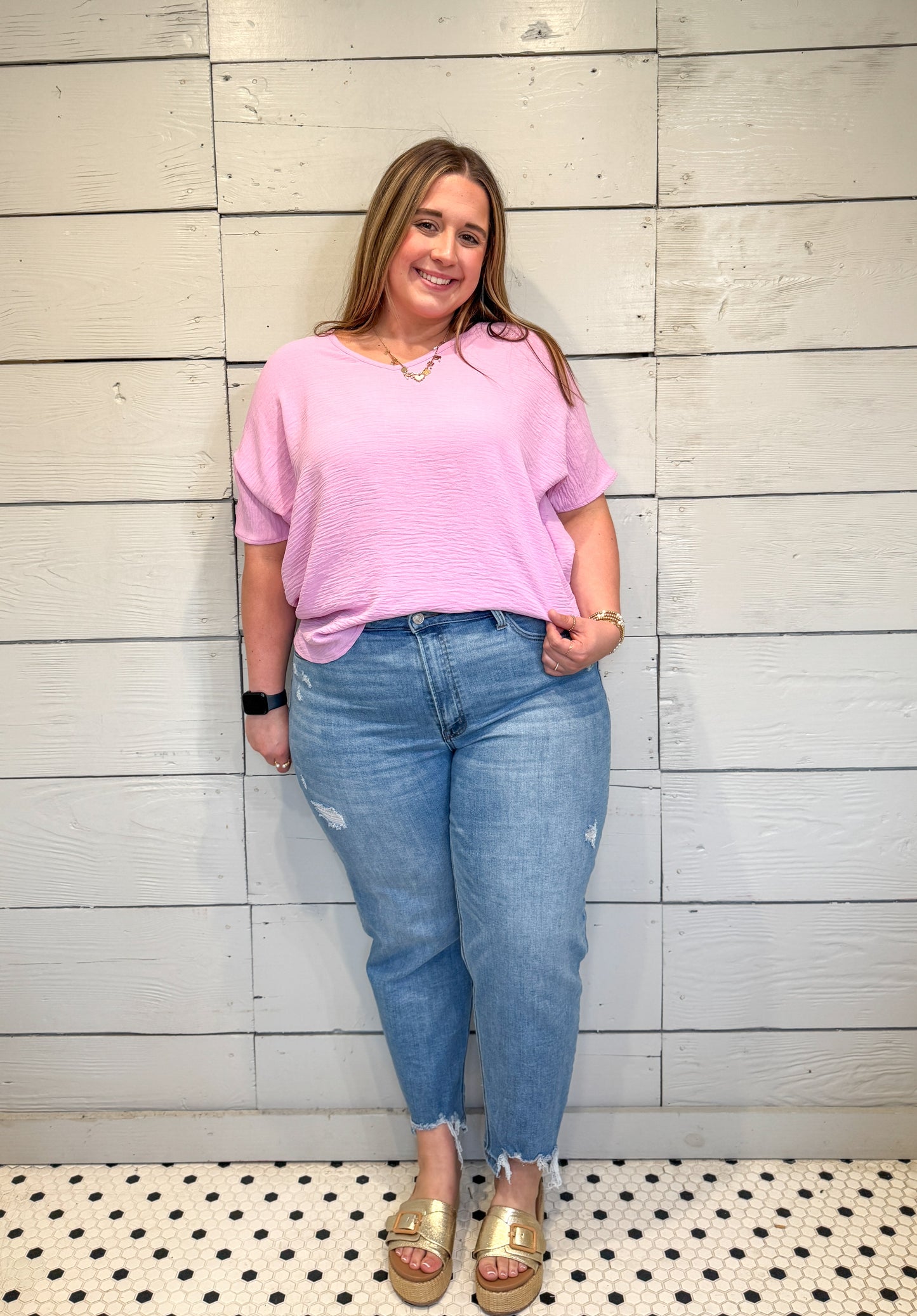 Woman wearing a pink top and blue jeans standing against a wooden wall.