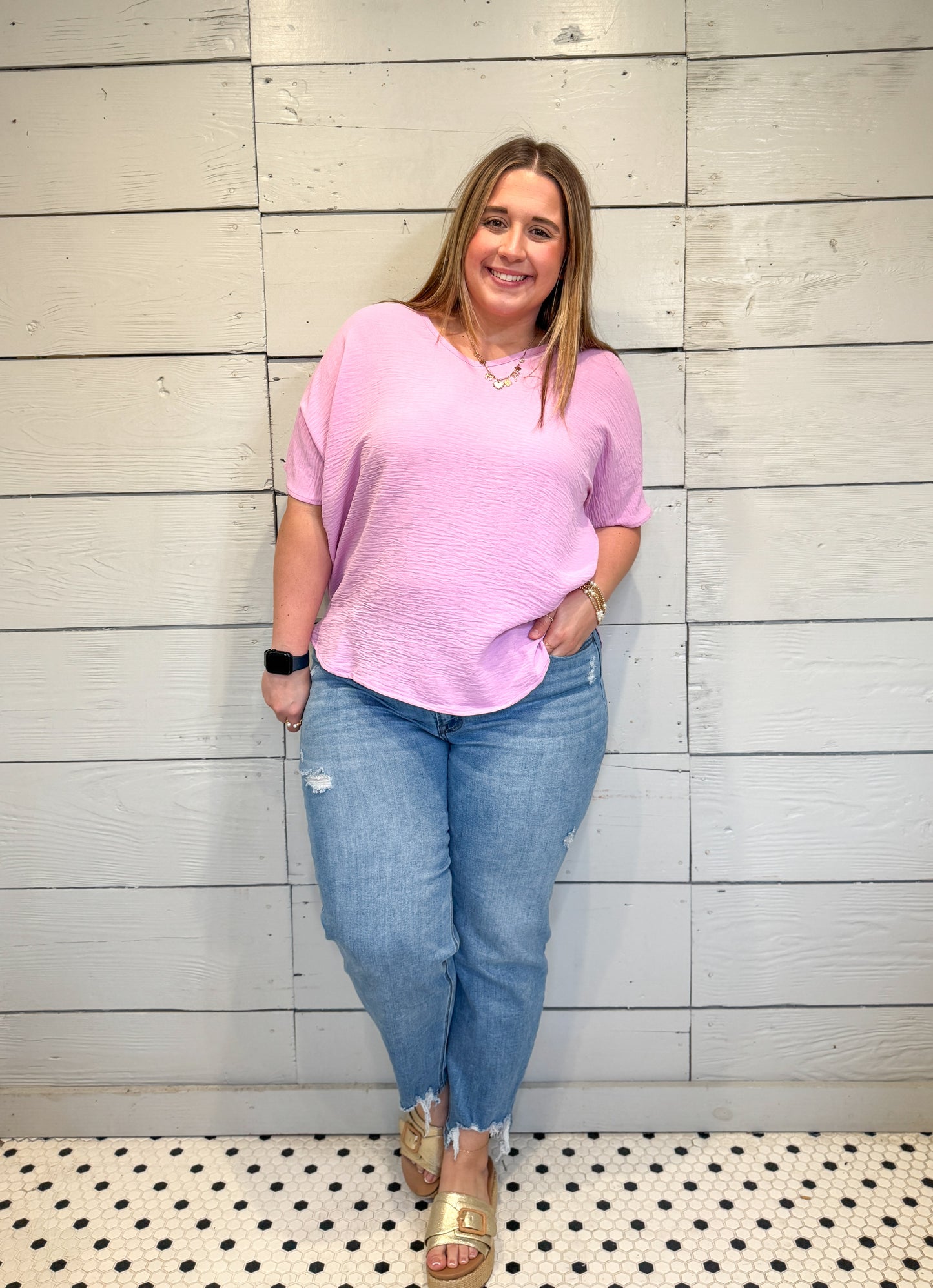 Woman wearing a pink top and blue jeans standing against a light wooden paneled wall.