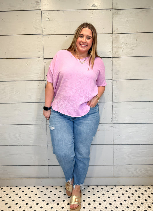 Woman wearing a pink top and blue jeans standing against a light wooden paneled wall.