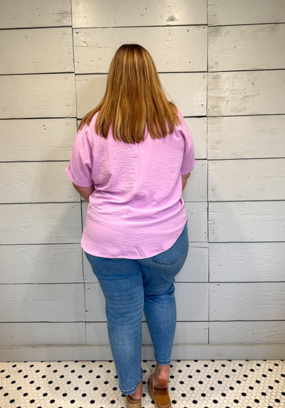 Person wearing a pink shirt and blue jeans standing against a light wooden paneled wall.