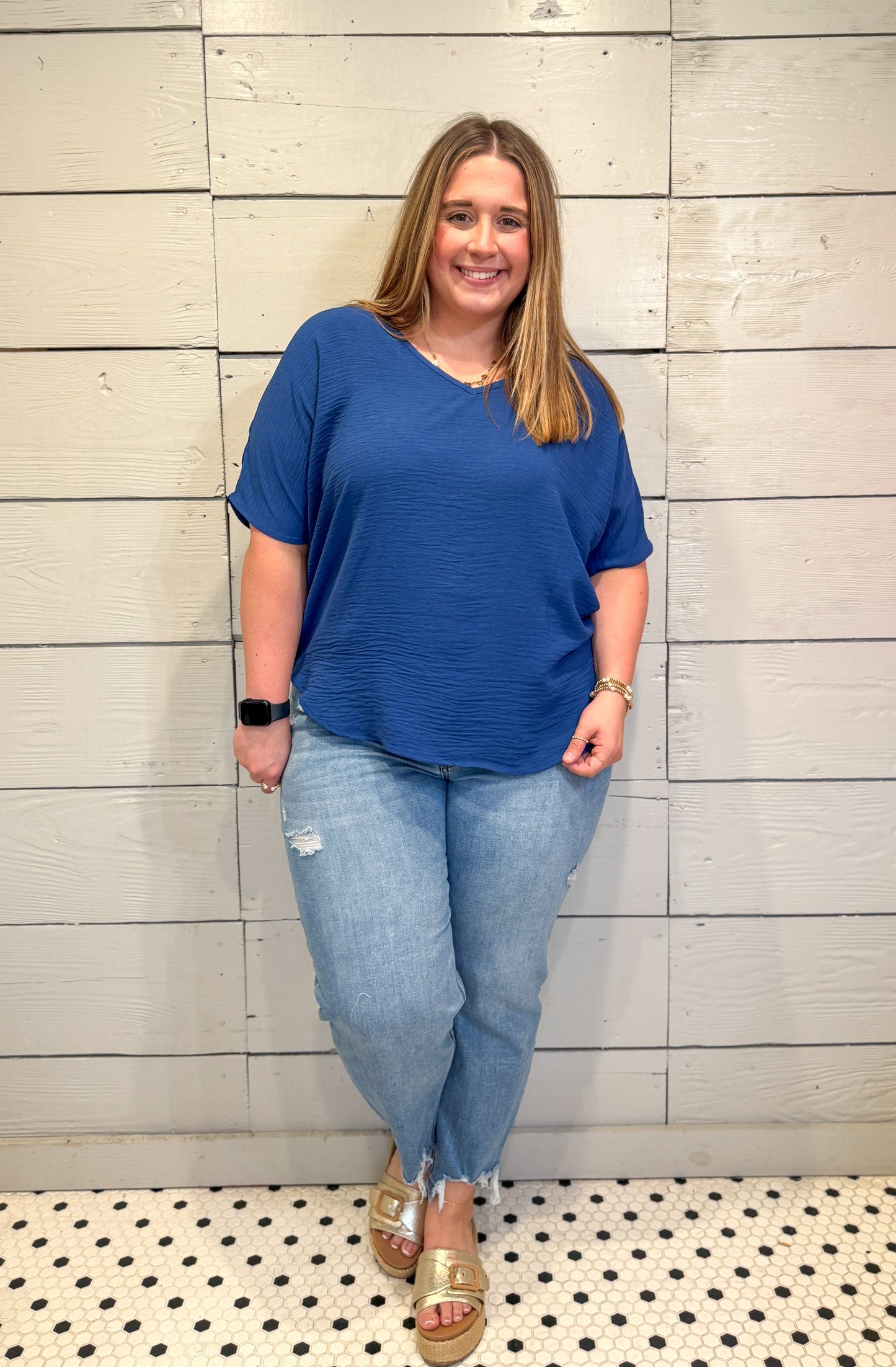 Woman wearing a blue top and jeans standing against a tiled wall.