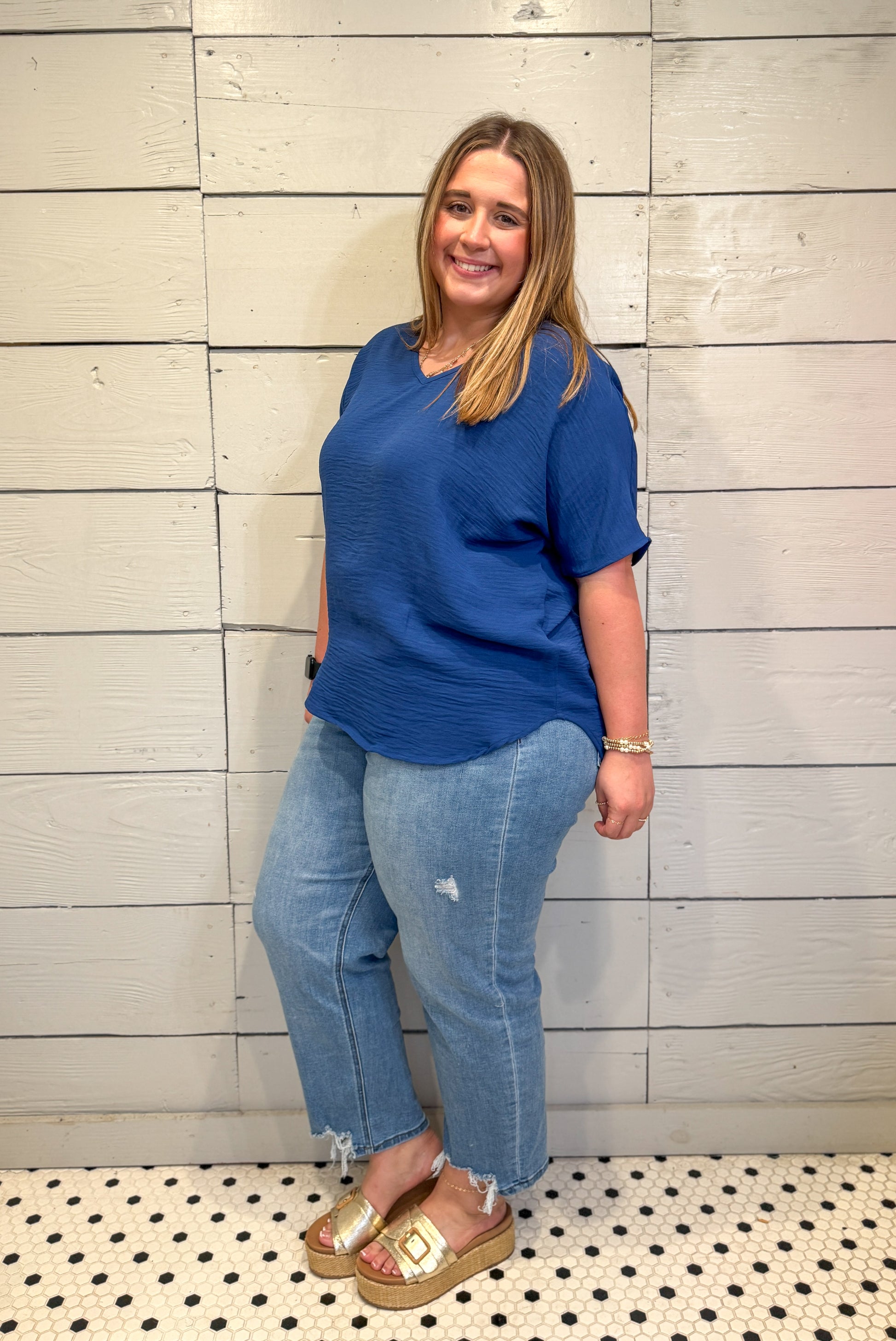 Woman wearing a blue shirt and jeans standing against a light wooden panel wall.