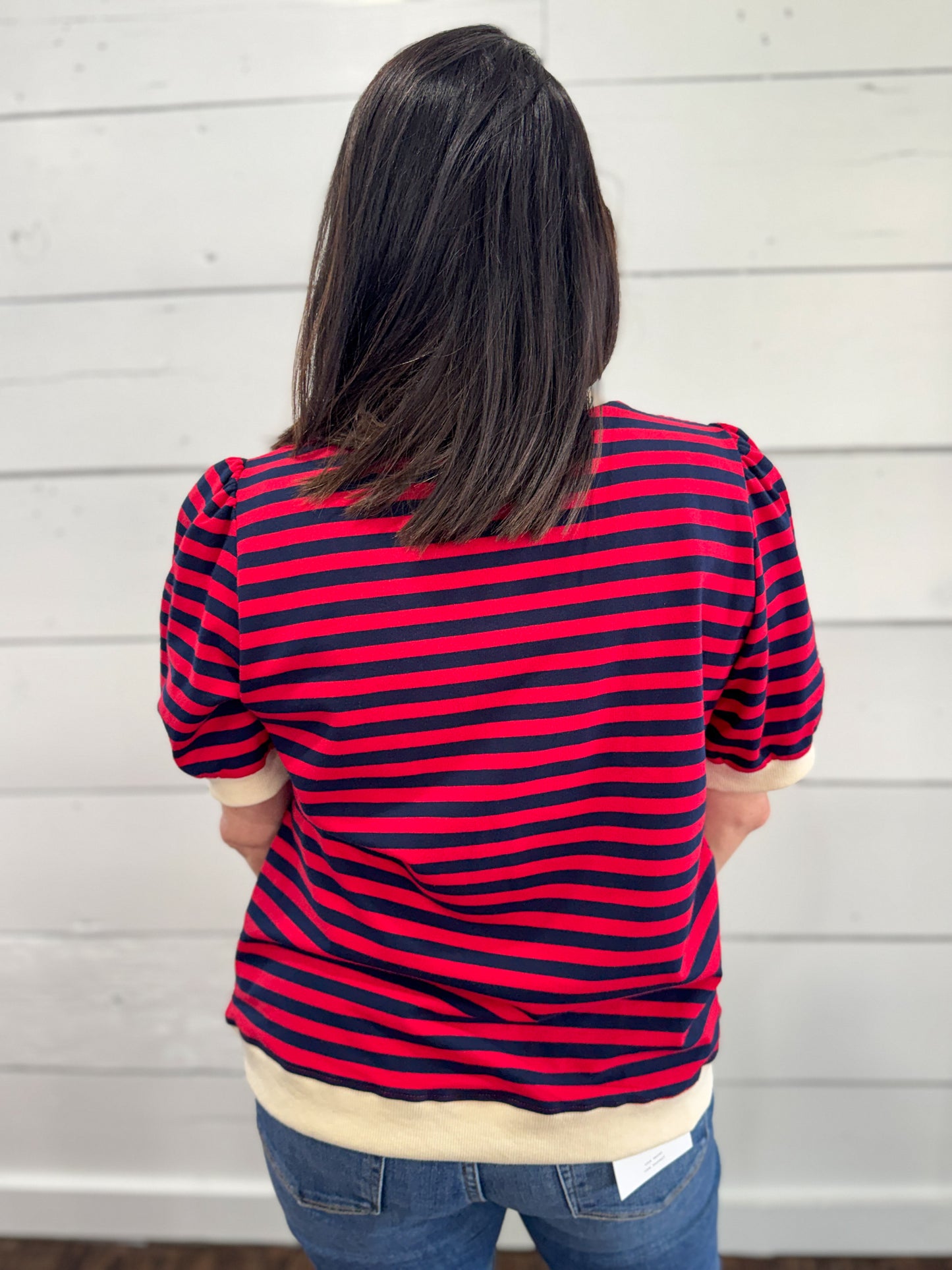 A woman viewed from behind wearing a red and navy blue horizontally striped short-sleeve top with cream ribbed trim, paired with blue jeans, standing against a white wood-paneled wall.
