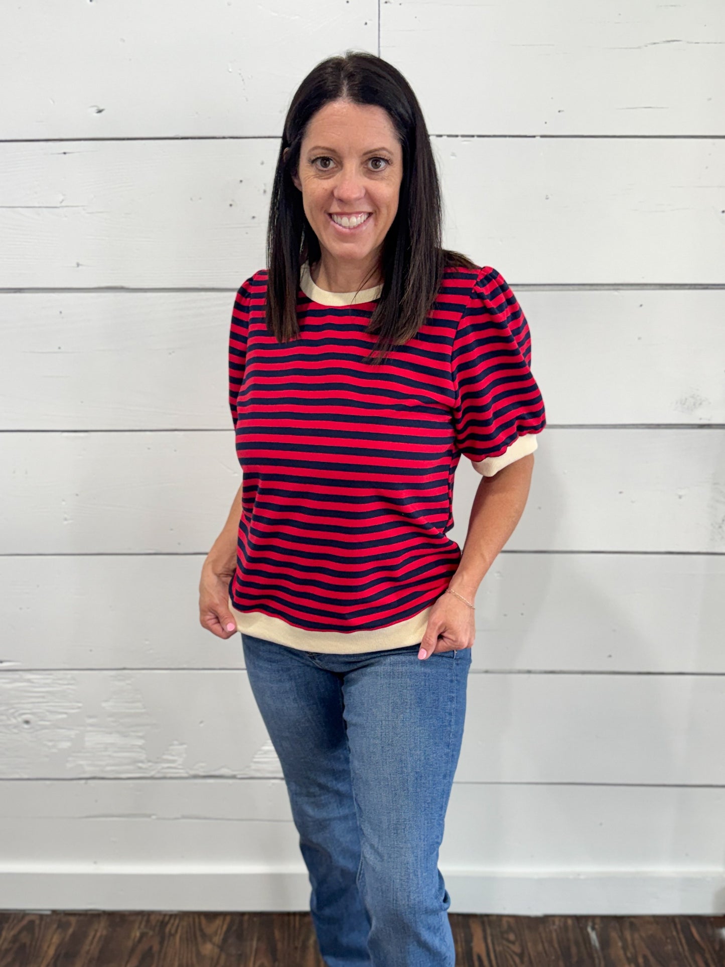 A woman wearing a red and navy blue horizontally striped short-sleeve t-shirt with cream ribbed trim, paired with blue jeans, standing against a white wood-paneled wall.