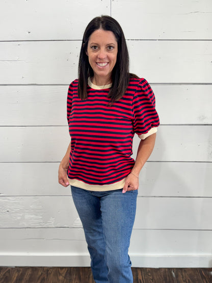 A woman wearing a red and navy blue horizontally striped short-sleeve t-shirt with cream ribbed trim, paired with blue jeans, standing against a white wood-paneled wall.