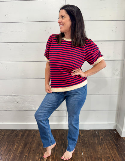A woman modeling a short-sleeve crewneck shirt with horizontal red and navy stripes and cream ribbed trim, paired with blue jeans, standing against a white shiplap wall.