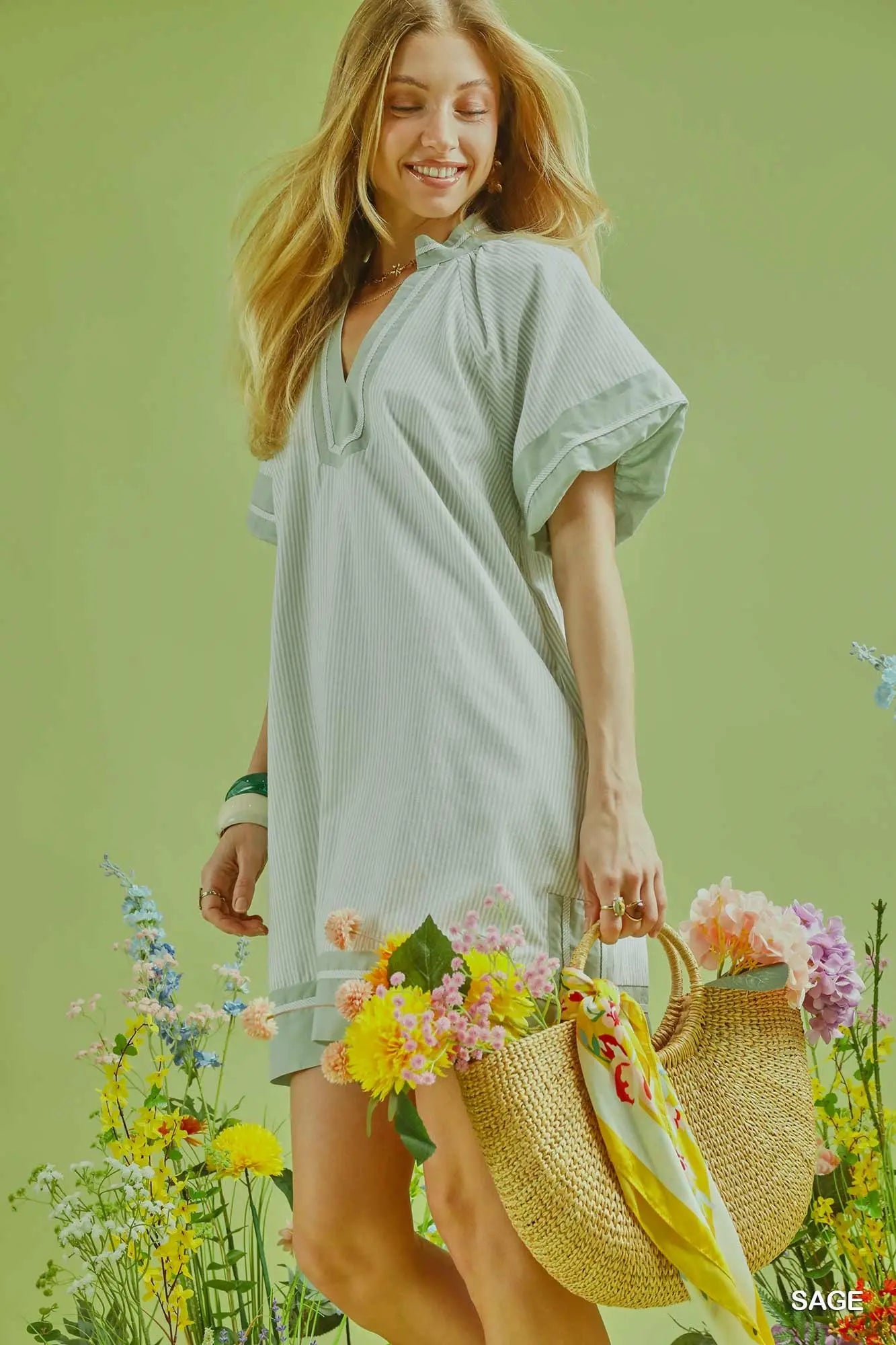 Woman in a light green dress holding flowers and a straw bag against a green background