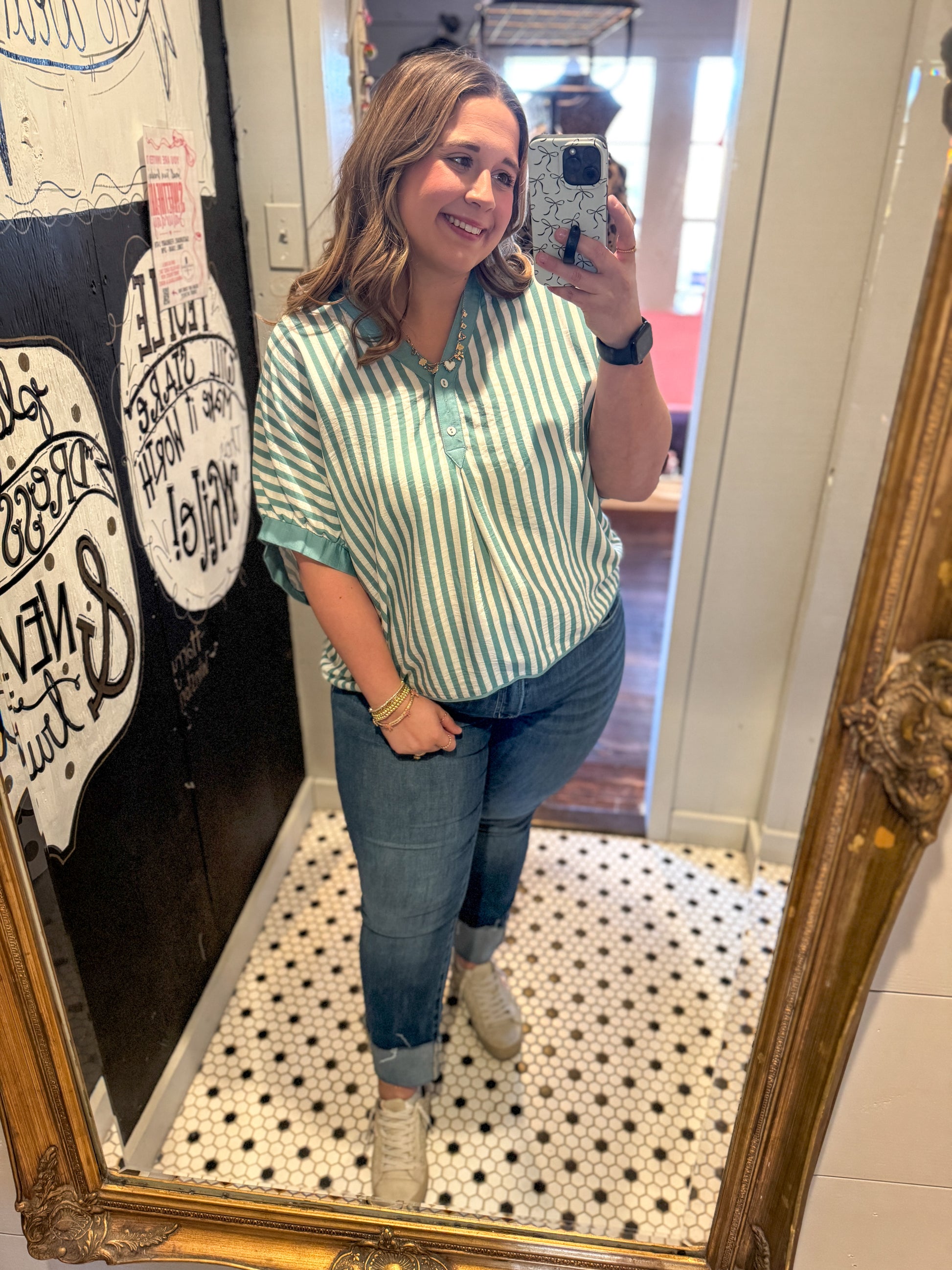 Woman taking a mirror selfie wearing a green and white striped shirt and blue jeans.
