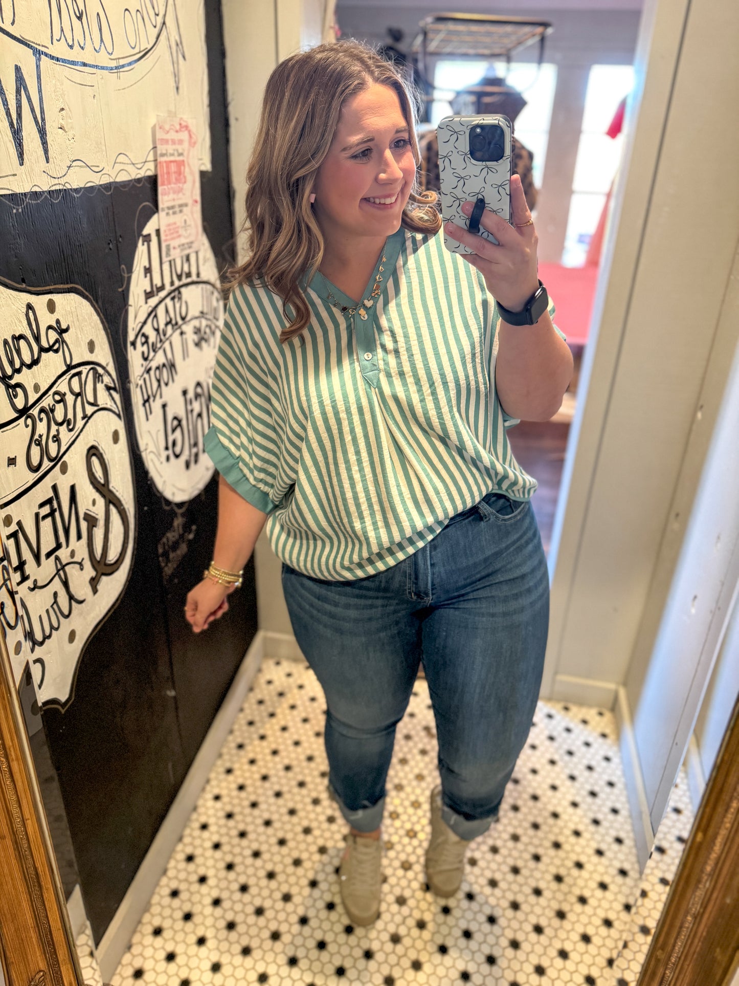 Woman taking a mirror selfie wearing a green and white striped shirt and blue jeans.