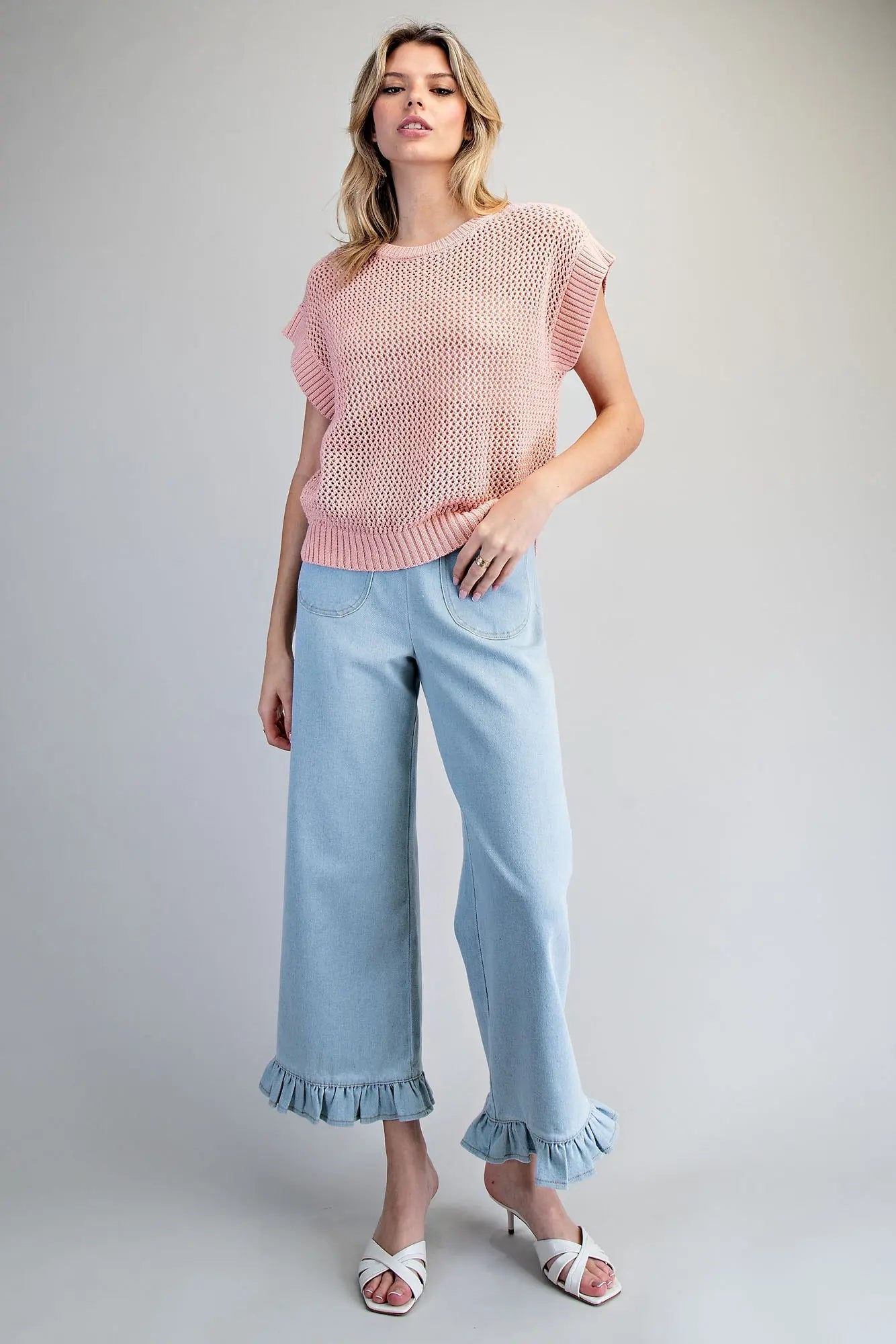 Woman wearing a pink sleeveless top and light blue jeans with ruffled cuffs on a gray background